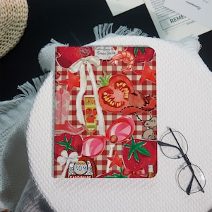 May include: A red and white checkered notebook with a whimsical design featuring tomatoes, cowboy boots, sunglasses, and a bottle of GRO tomato sauce. The notebook is decorated with various red accents.