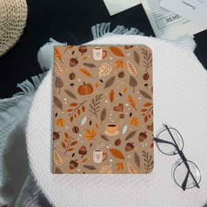 May include: A tan notebook with a fall-themed pattern featuring leaves, acorns, coffee cups, and a pumpkin. The design includes shades of brown, orange, and white. The notebook is rectangular and appears to be closed.
