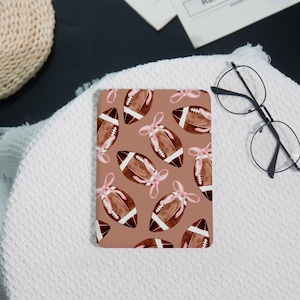 May include: A brown notebook with a repeating pattern of watercolor footballs tied with pink bows. The background is a solid, muted brown color. The notebook is on a white textured surface.