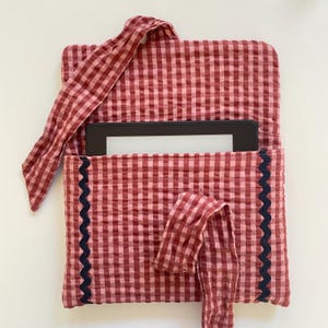 Puede incluir: Una funda para lector de libros electr&oacute;nicos de tela de cuadros rojos y rosas con un ribete festoneado azul marino oscuro. La funda tiene un cierre de lazo de tela a juego y un asa. Un lector de libros electr&oacute;nicos es visible en el interior.
