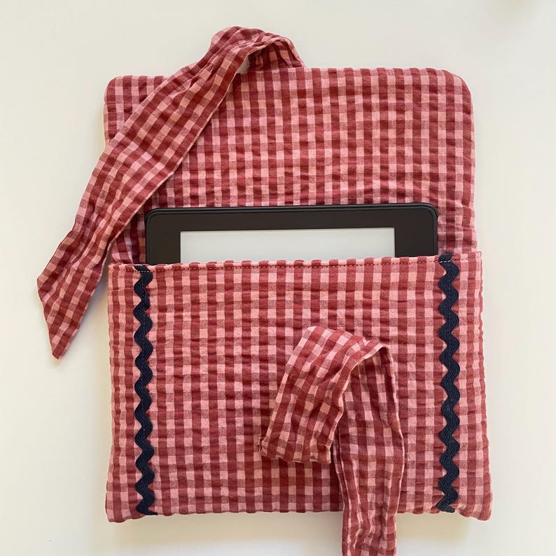 Puede incluir: Una funda para lector de libros electr&oacute;nicos de tela de cuadros rojos y rosas con un ribete festoneado azul marino oscuro. La funda tiene un cierre de lazo de tela a juego y un asa. Un lector de libros electr&oacute;nicos es visible en el interior.