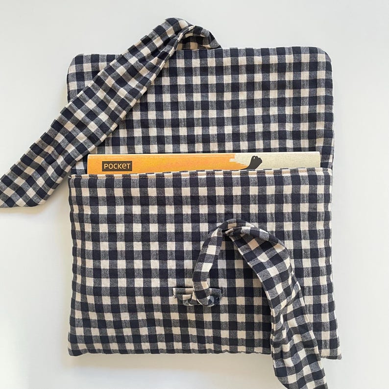 Puede incluir: Una funda para libros con estampado de cuadros vichy en blanco y negro con cierre de lazo a juego. La funda tiene un bolsillo con la palabra "pocket" impresa en un libro en su interior. Dise&ntilde;ada para proteger libros.