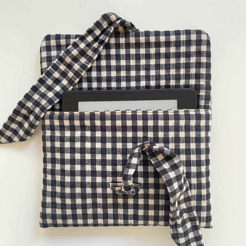 Puede incluir: Una funda para tableta con estampado de cuadros vichy en blanco y negro con un lector de libros electr&oacute;nicos negro en el interior. La funda tiene un cierre de solapa con una corbata de tela y un asa de tela. La tela es un estampado de cuadros cl&aacute;sico.