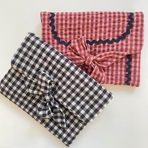 Puede incluir: Dos bolsas de tela de cuadros vichy. Una es azul marino y blanca, la otra roja y blanca. Ambas tienen un cierre de lazo. La bolsa roja y blanca tiene un ribete festoneado azul marino. Estas bolsas son ideales para guardar objetos peque&ntilde;os.