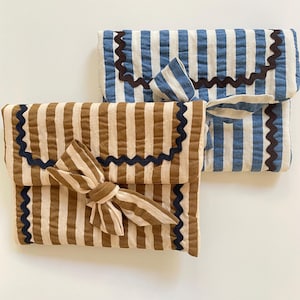 Pode incluir: Duas clutches de tecido listrado. Uma é marrom e creme com uma borda ondulada azul escuro e laço. A outra é azul e branca com uma borda ondulada marrom e um laço pequeno. Ambas são retangulares.