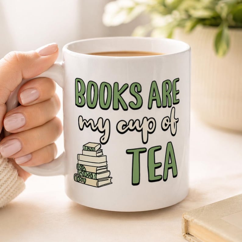 May include: White ceramic mug with the text "BOOKS ARE my cup of TEA" in green and black lettering. A stack of books is illustrated below the text. The mug is filled with a brown liquid.