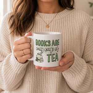 May include: White ceramic mug with the text "BOOKS ARE my cup of TEA" in green and black lettering. The mug also features a small stack of books. The mug is held by a person wearing a cream-colored sweater.
