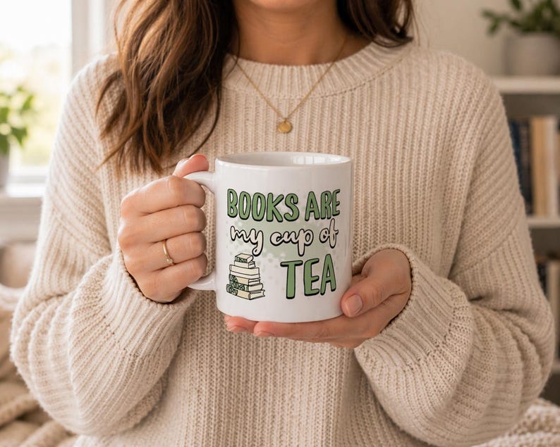 May include: White ceramic mug with the text "BOOKS ARE my cup of TEA" in green and black lettering. The mug also features a small stack of books. The mug is held by a person wearing a cream-colored sweater.