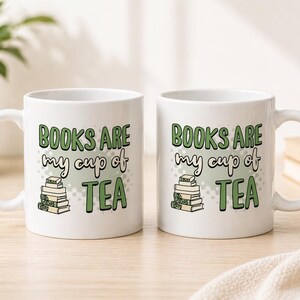 May include: Two white ceramic mugs with the text "BOOKS ARE my cup of TEA" in green and black lettering. The mugs feature a stack of books graphic. The mugs are on a light-colored wooden surface.