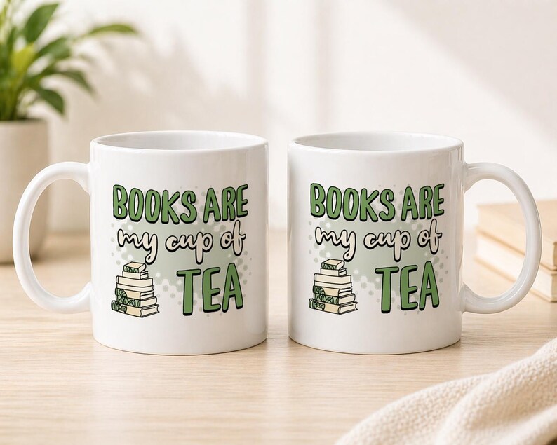 May include: Two white ceramic mugs with the text "BOOKS ARE my cup of TEA" in green and black lettering. The mugs feature a stack of books graphic. The mugs are on a light-colored wooden surface.