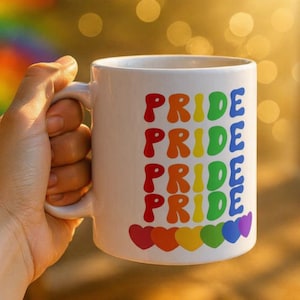 Puede incluir: Taza de cer&aacute;mica blanca con la palabra "PRIDE" repetida en colores del arco&iacute;ris. La taza tiene un asa y una fila de corazones de colores del arco&iacute;ris en la parte inferior. La taza est&aacute; sostenida por una persona.