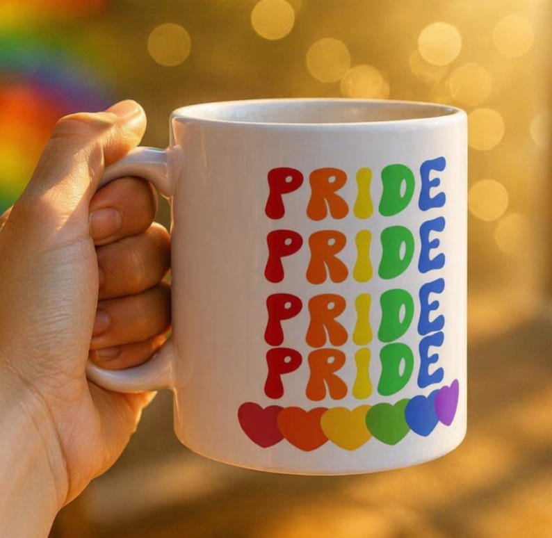 Puede incluir: Taza de cer&aacute;mica blanca con la palabra "PRIDE" repetida en colores del arco&iacute;ris. La taza tiene un asa y una fila de corazones de colores del arco&iacute;ris en la parte inferior. La taza est&aacute; sostenida por una persona.