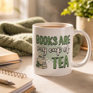 May include: White ceramic mug with the text "BOOKS ARE my cup of TEA" in green lettering. A stack of books is illustrated below the text. The mug is filled with a hot beverage, and steam rises from the top. A cozy reading theme.
