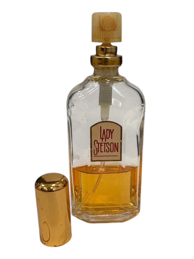 Peut inclure: Un flacon de parfum en verre transparent avec un bouchon dor&eacute; et un couvercle dor&eacute; s&eacute;par&eacute;. Le flacon porte une &eacute;tiquette avec l'inscription "LADY STETSON" dans un cadre rectangulaire. Le parfum &agrave; l'int&eacute;rieur est de couleur ambre.