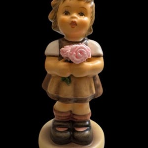 以下が含まれることがあります： ピンクのバラの花束を持った少女の陶器人形。少女は茶色のドレス、白いブラウス、黒い靴を履いています。人形は丸いベージュ色の台座の上に立っています。