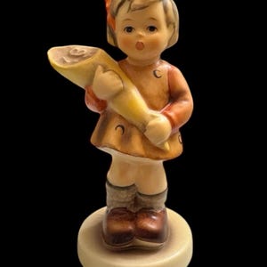 Puede incluir: Una figura de cerámica de una niña pequeña sosteniendo un objeto amarillo grande, posiblemente un cuerno o un pergamino. Lleva un vestido marrón con mangas naranjas y un lazo rojo en el pelo. La figura está sobre una base de color crema.
