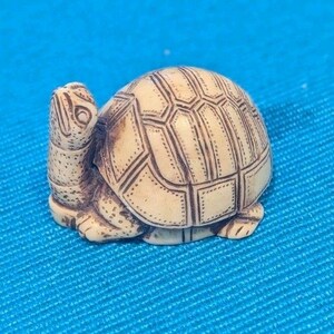 Op de afbeelding: Een kleine, gedetailleerde schildpadfiguur met een crèmekleurig lichaam en een patroon op de schild. De schildpadschild heeft een geometrisch ontwerp met donkerbruine lijnen. De figuur staat op een blauwe achtergrond.
