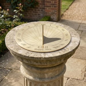 May include: A circular brass sundial featuring Roman numerals from I to XII, a central vertical gnomon, and the engraved text E.D.H and Happy 50th. The sundial is mounted on a weathered, cylindrical stone pedestal with a fluted base design.