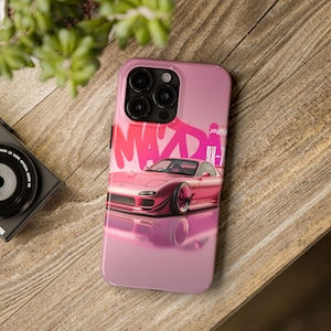Könnte beinhalten: Rosa Handyhülle mit einem stilisierten Bild eines rosa Mazda RX-7 Sportwagens. Die Hülle hat ein glänzendes Finish und die Wörter "MAZD" und "RX-7" sind in einer Graffiti-Schriftart zu sehen. Die Hülle liegt auf einer Holzoberfläche.