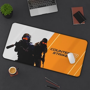 May include: A rectangular mouse pad featuring a Counter-Strike design. The pad has a white and orange background with two silhouetted figures holding weapons. The words "COUNTER STRIKE" are printed on the pad.