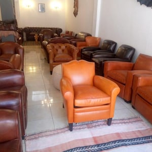 Puede incluir: Una sala de exposición con varios sillones de cuero en tonos marrones y naranjas. Las sillas tienen diferentes diseños, incluyendo respaldos con botones y brazos enrollados. Una alfombra estampada está en primer plano.