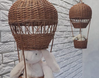 Op de afbeelding: Twee hangende decoraties van rieten luchtballonnen. Elke ballon heeft een geweven mand en een pluchen konijn erin. De ballonnen hangen aan het plafond tegen een witte bakstenen muur. De ballonnen zijn bruin en de konijnen zijn wit.