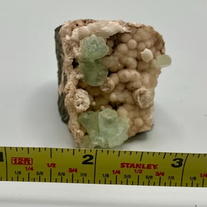 Op de afbeelding: Een close-up van een kleine steen met groene kristallen ingebed in een witte matrix. De steen is ongeveer 5 cm lang en wordt gemeten met een meetlint.