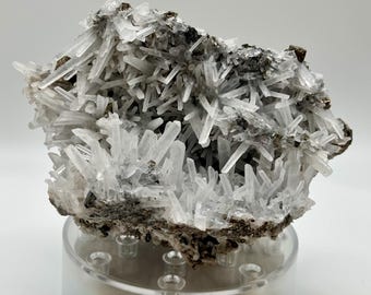 Może przedstawiać: Gromada przezroczystych kryształów kwarcu z szarą, metaliczną podstawą. Kryształy są ostre i spiczaste, a ich układ jest przypadkowy. Podstawa jest szorstka i fakturowana. Gromada ma około 12,5 cm szerokości.