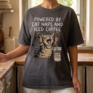 May include: A charcoal gray short-sleeve t-shirt featuring a graphic of a yawning tabby cat with its paws raised. The text reads, "POWERED BY CAT NAPS AND ICED COFFEE," and the cat is holding a cup labeled "CAT NAP FUEL."
