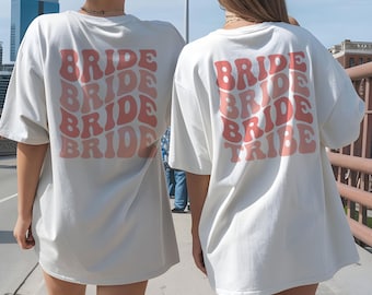 May include: Two oversized white t-shirts with the word "BRIDE" stacked in a retro font on the back. One shirt also says "TRIBE". The text is in shades of pink and orange. The shirts are worn by two people outdoors.