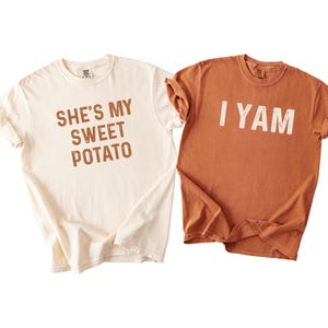 May include: Two matching t-shirts, one cream-colored and one rust-colored. The cream shirt has the text "SHE'S MY SWEET POTATO" in brown. The rust shirt has the text "I YAM" in white. Both shirts have rolled-up sleeves.