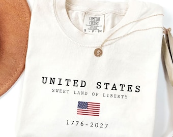 May include: An off-white t-shirt featuring the black text UNITED STATES, SWEET LAND OF LIBERTY, and 1776-2027, with a small American flag graphic in the center. The shirt has a Comfort Colors brand label at the neck and a gold-toned necklace.