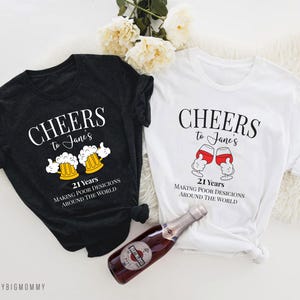 May include: Two black and white t-shirts with a humorous 21st birthday design. The shirts feature the text "Cheers to Jane's 21 Years Making Poor Decisions Around the World" with a graphic of a beer mug or wine glass.