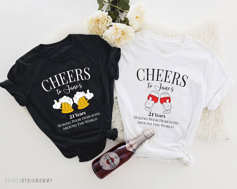 May include: Two black and white t-shirts with a humorous 21st birthday design. The shirts feature the text "Cheers to Jane's 21 Years Making Poor Decisions Around the World" with a graphic of a beer mug or wine glass.