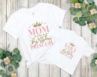 May include: Two white t-shirts with pink and gold lettering. The larger shirt says "Mom of the Birthday Princess" with a gold crown. The smaller shirt says "Sara the Birthday Princess" with a gold crown.