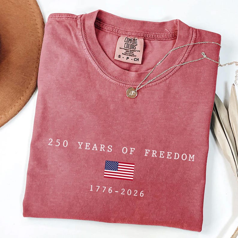 K&ouml;nnte beinhalten: Ein staubrosa Comfort Colors T-Shirt mit dem Text "250 YEARS OF FREEDOM" und der amerikanischen Flagge. Das Shirt hat auch die Daten "1776-2026" aufgedruckt. Eine silberne Halskette mit Anh&auml;nger liegt auf dem Shirt.