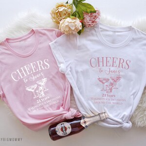 Peut inclure: Deux t-shirts roses et blancs avec le texte "Cheers to Jane's 21 Years Making Pour Decisions Around The World" et un graphique de verres &agrave; martini.