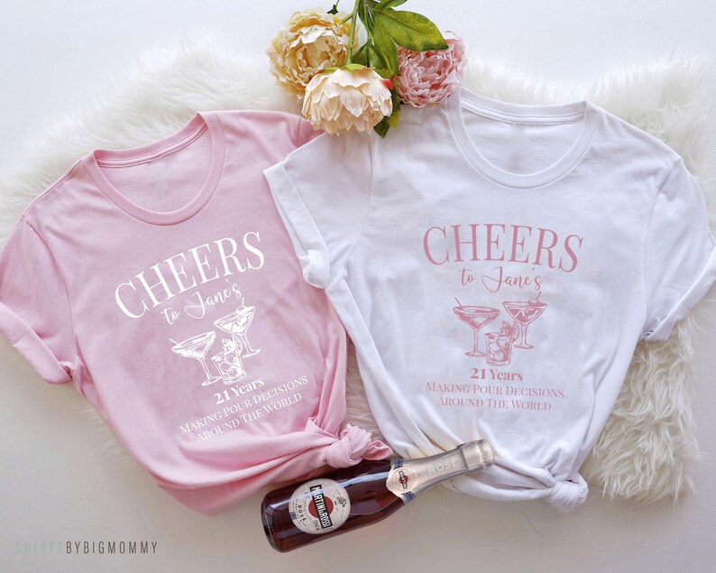 Peut inclure: Deux t-shirts roses et blancs avec le texte "Cheers to Jane's 21 Years Making Pour Decisions Around The World" et un graphique de verres &agrave; martini.
