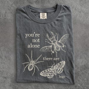 May include: A charcoal gray t-shirt featuring a white line-art graphic of a stag beetle, a spider, and a moth. The text "you're not alone there are bugs" is printed in a serif font. The shirt has a crew neckline and a Comfort Colors brand label.