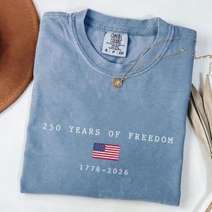 K&ouml;nnte beinhalten: Ein blaues Comfort Colors T-Shirt mit dem Text "250 YEARS OF FREEDOM" und einer amerikanischen Flaggen-Grafik. Das Shirt zeigt auch die Jahreszahlen "1776-2026". Eine goldene Halskette mit Anh&auml;nger liegt auf dem Shirt.