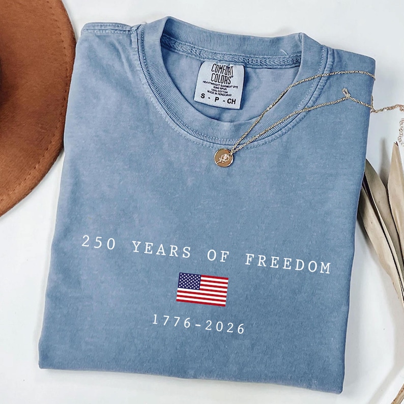 K&ouml;nnte beinhalten: Ein blaues Comfort Colors T-Shirt mit dem Text "250 YEARS OF FREEDOM" und einer amerikanischen Flaggen-Grafik. Das Shirt zeigt auch die Jahreszahlen "1776-2026". Eine goldene Halskette mit Anh&auml;nger liegt auf dem Shirt.