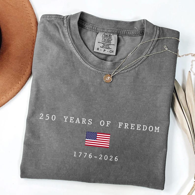 K&ouml;nnte beinhalten: Graues T-Shirt mit dem Text "250 YEARS OF FREEDOM" und der amerikanischen Flagge. Die Daten "1776-2026" sind darunter aufgedruckt. Eine silberne Halskette mit Anh&auml;nger liegt auf dem Shirt.