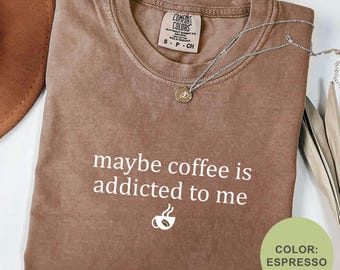 May include: Espresso-colored t-shirt with the phrase "maybe coffee is addicted to me" and a coffee cup graphic. The shirt has a crew neck and short sleeves. A silver necklace with a pendant is draped across the shirt.
