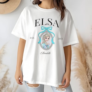 May include: White t-shirt featuring the name "ELSA" in black, above a portrait of a character in a blue oval frame with a blue bow. The text "EST. 2013" and "Arendelle" are also printed on the shirt.