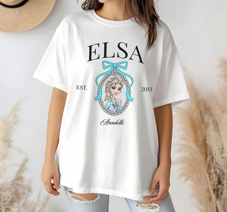 May include: White t-shirt featuring the name "ELSA" in black, above a portrait of a character in a blue oval frame with a blue bow. The text "EST. 2013" and "Arendelle" are also printed on the shirt.