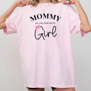 May include: A light pink t-shirt with the text "MOMMY of the BIRTHDAY Girl" in black script. A small pink bow is above the word "Girl". The shirt has short sleeves and a relaxed fit.