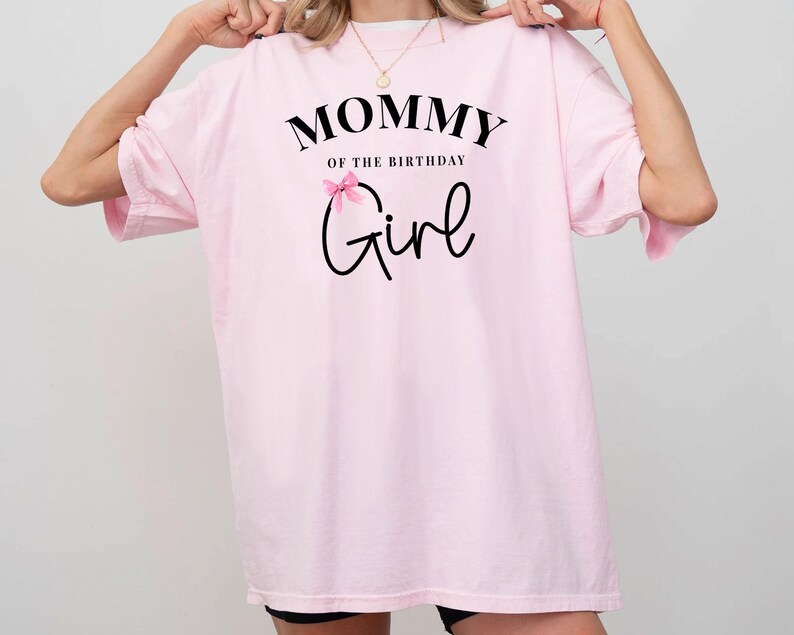 May include: A light pink t-shirt with the text "MOMMY of the BIRTHDAY Girl" in black script. A small pink bow is above the word "Girl". The shirt has short sleeves and a relaxed fit.