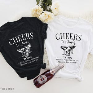 Peut inclure: Deux t-shirts noirs et blancs avec le texte "Cheers to Jane's 21 Years Making Pour Decisions Around the World" et un graphique de verres &agrave; martini.