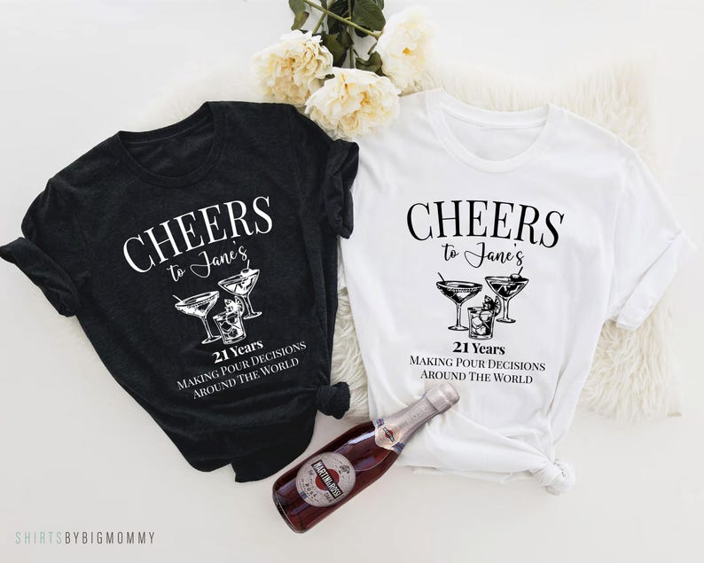 Peut inclure: Deux t-shirts noirs et blancs avec le texte "Cheers to Jane's 21 Years Making Pour Decisions Around the World" et un graphique de verres &agrave; martini.