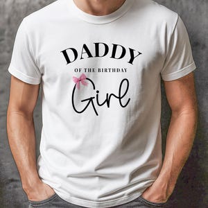 May include: White t-shirt with the text "DADDY of the BIRTHDAY Girl" in black script. A pink bow is above the word "Girl". The shirt is a crew neck and has short sleeves.